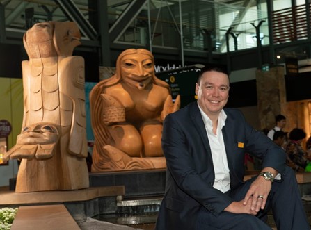 Larry Shuter, Indigenous man wearing a dark blue suit, sitting to the right of two indigenous style carved wood sculptures 