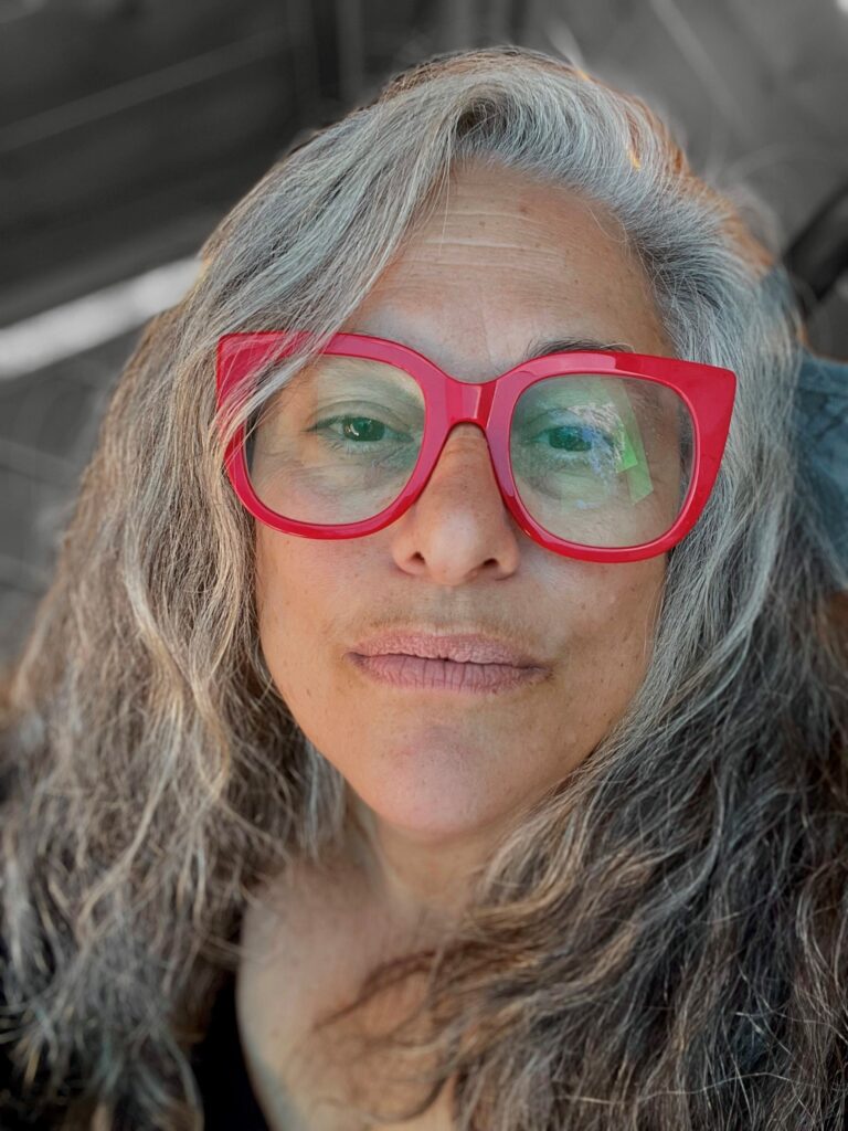 Woman with light skin and long gray hair wearing big red glasses and black shirt.