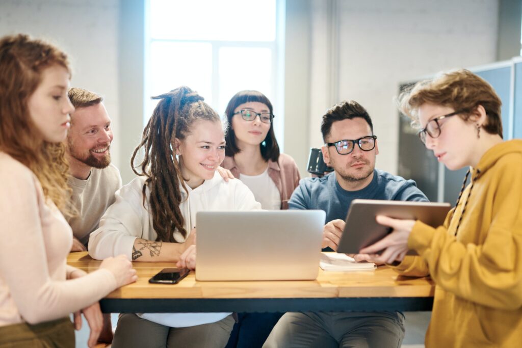 An image of six colleagues gathered around a laptop.