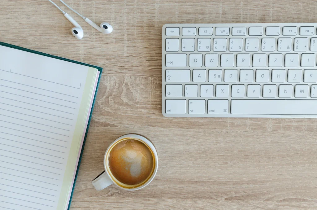 Image d'une tassse de café, d'un carnet de notes, d'écouteurs et d'un clavier sur un bureau en bois.